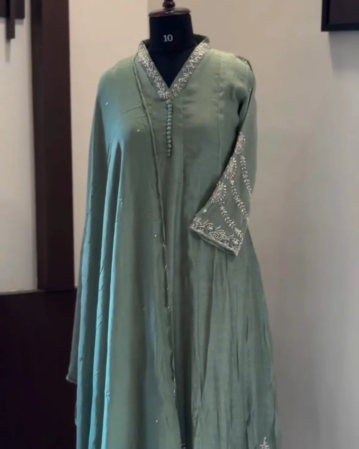 A sage green long kurta displayed on a black mannequin torso, featuring silver mirror work embroidery on the V-neckline and sleeve cuff.