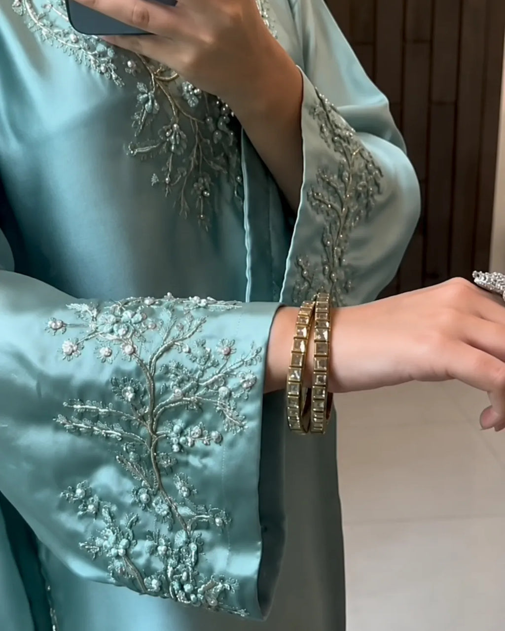 A close-up of a person wearing a light blue satin garment with intricate silver floral embroidery on the sleeve, paired with stacked gold bangles on the wrist.