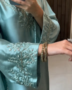 A close-up of a person wearing a light blue satin garment with intricate silver floral embroidery on the sleeve, paired with stacked gold bangles on the wrist.