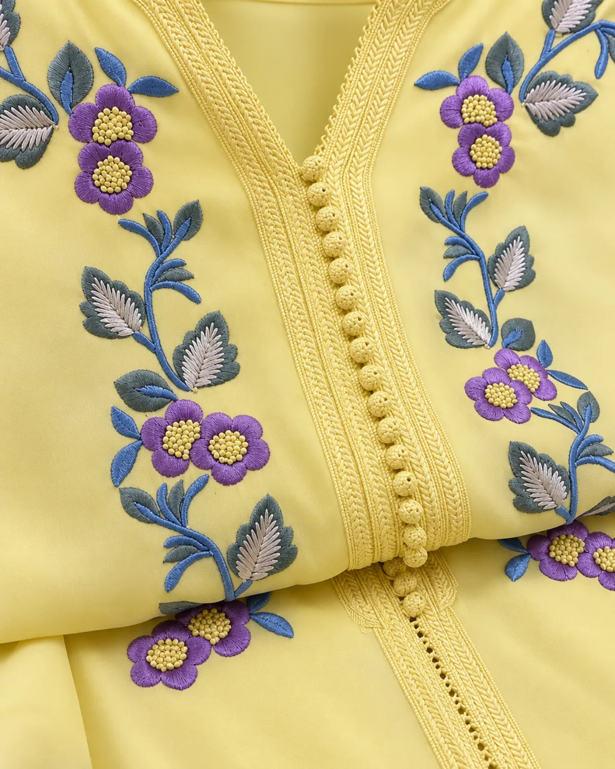 Close-up of a pale yellow garment with embroidered floral vines in purple, blue, green, and gray, plus a braided trim running down the center front.
