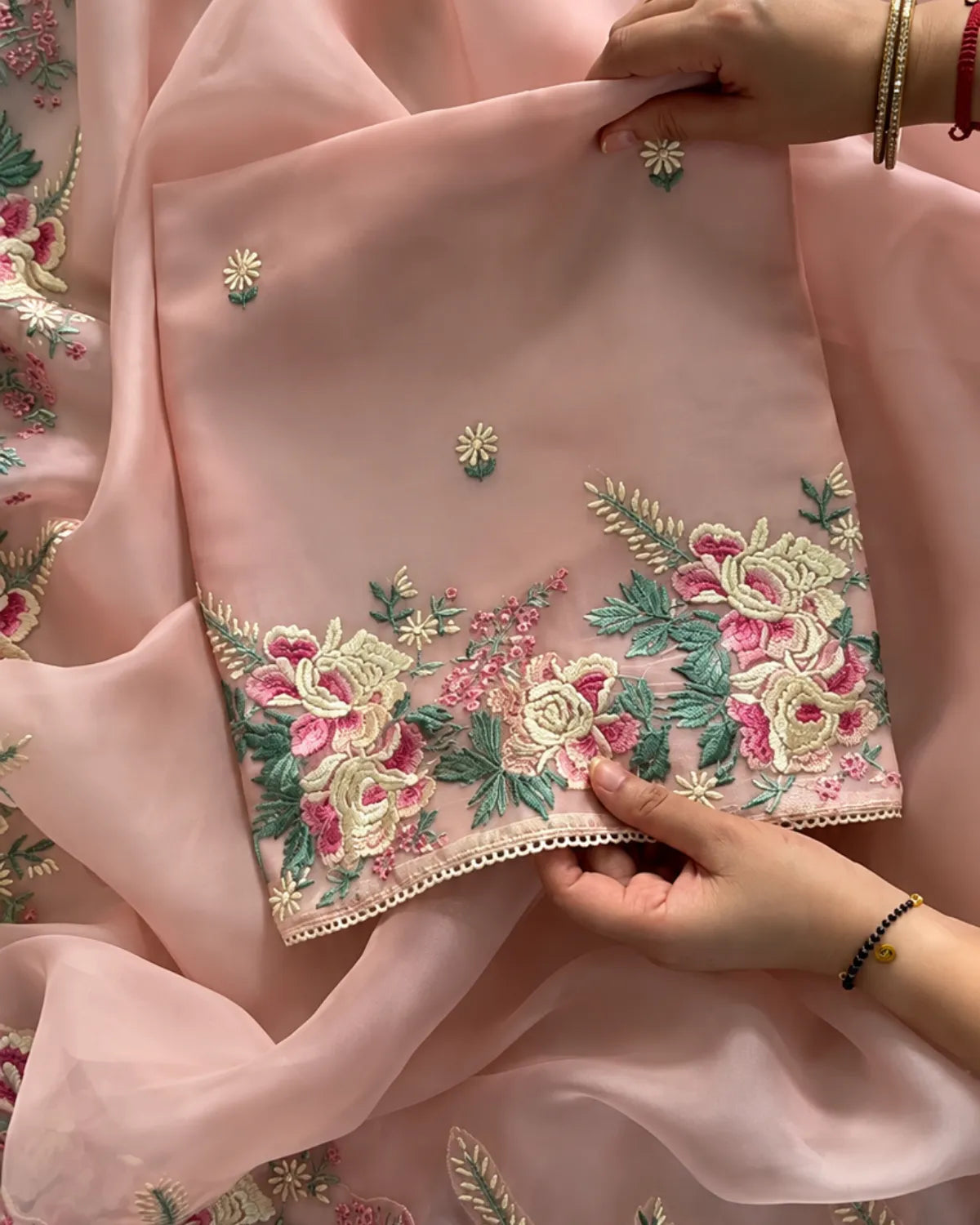 A close-up of a light pink embroidered fabric, likely a dupatta or scarf, held by hands to showcase its detailed floral embroidery. The fabric features vibrant pink and cream flowers with green leaves and small embroidered motifs, finished with a decorative scalloped border.
