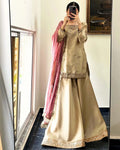 A woman takes a mirror selfie wearing a traditional beige and gold embroidered kurta set with a pink dupatta draped over her shoulder, set against an indoor background.