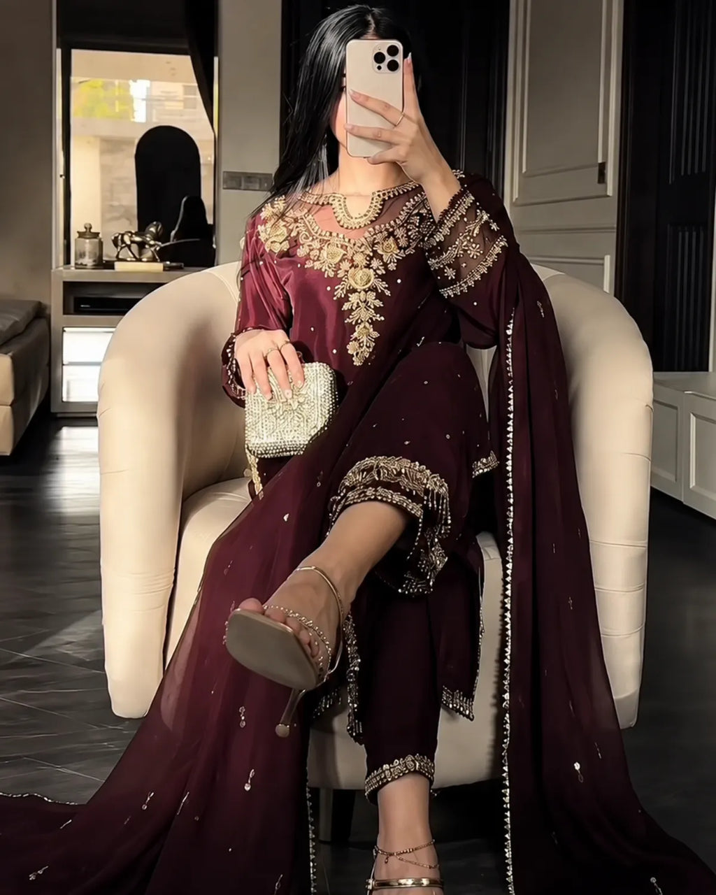 A woman takes a mirror selfie wearing a deep maroon traditional outfit with heavy gold embroidery, seated in a cream-colored armchair. The outfit includes a long-sleeved top, matching pants, and a draped dupatta.
