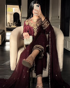 A woman takes a mirror selfie wearing a deep maroon traditional outfit with heavy gold embroidery, seated in a cream-colored armchair. The outfit includes a long-sleeved top, matching pants, and a draped dupatta.