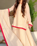 A woman models a cream-colored three-piece salwar kameez suit with vibrant red lace trim. The set includes a long-sleeved tunic, matching wide-leg trousers, and a coordinating dupatta draped over her arm.