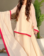 A woman models a cream-colored three-piece salwar kameez suit with vibrant red lace trim. The set includes a long-sleeved tunic, matching wide-leg trousers, and a coordinating dupatta draped over her arm.