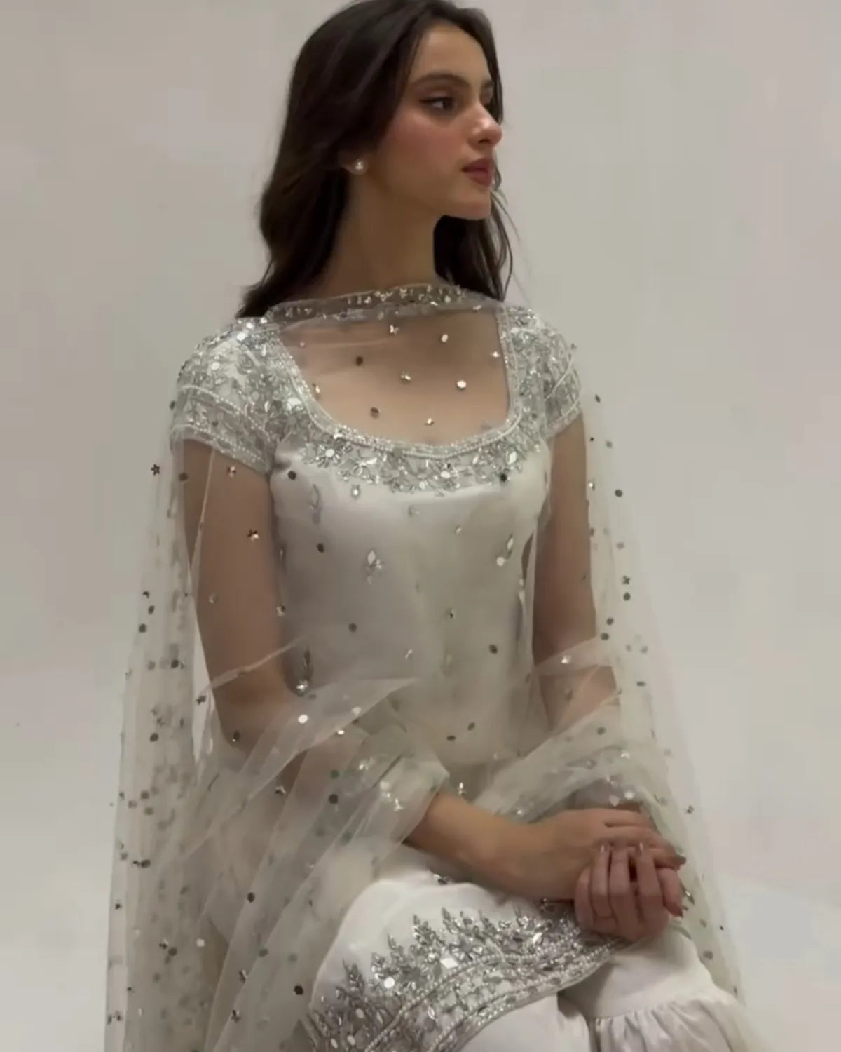 A woman wearing a white embellished traditional outfit with a sheer silver-embroidered dupatta, seated against a plain light background.