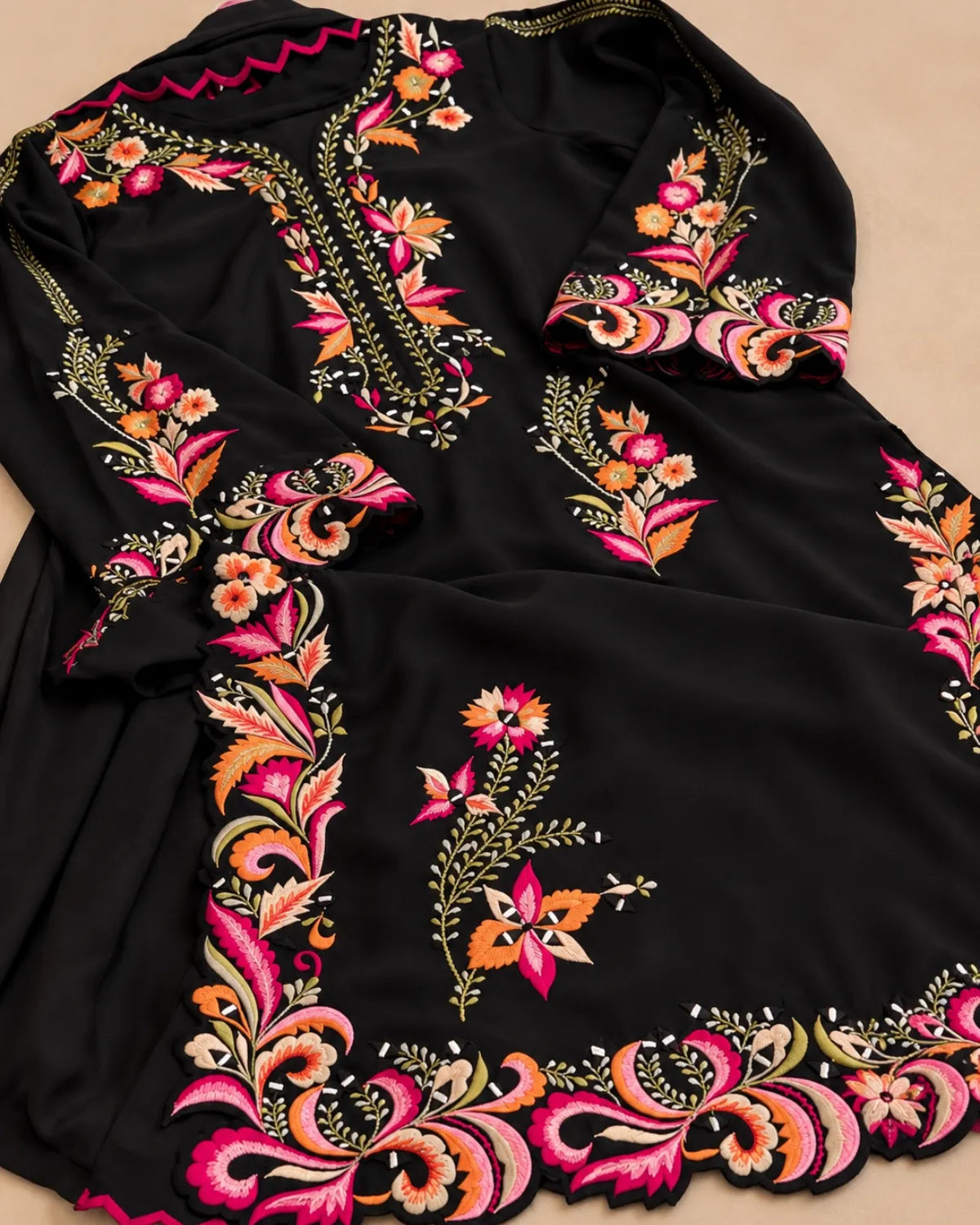 A black embroidered tunic or kurta laid flat on a beige background, featuring vibrant floral and paisley embroidery in pink, orange, yellow, and green.