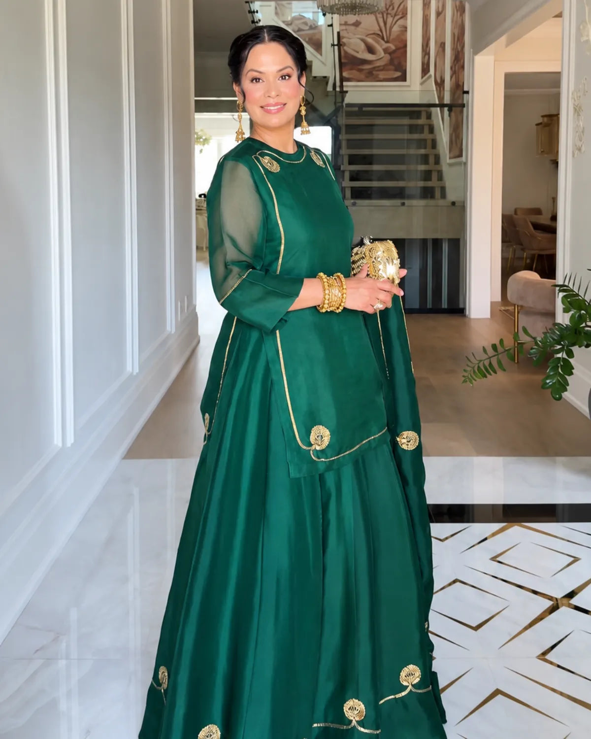 A woman wearing a dark green traditional dress with gold embroidery and sheer sleeves, holding a gold clutch purse in a modern indoor hallway.