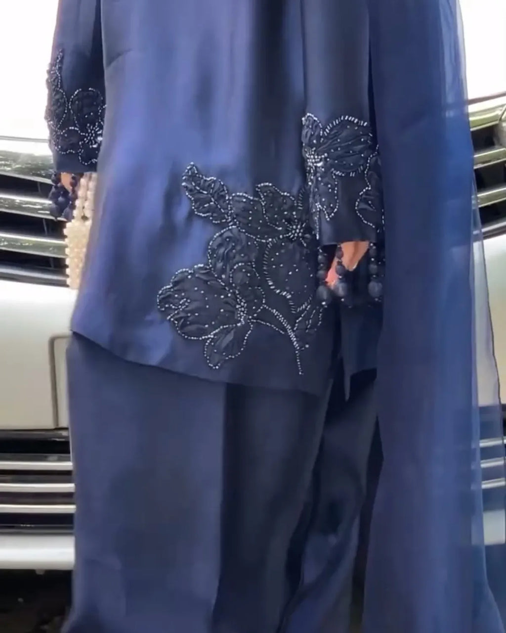 A close-up of a navy blue traditional outfit with intricate silver beaded floral embroidery on the sleeves and lower tunic, worn by a model standing in front of a car.