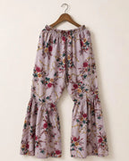 A pair of wide-leg, tiered pants with an elastic waistband, featuring a dusty lavender floral print with red, green, and gold details, hanging on a wooden hanger against a plain beige wall.