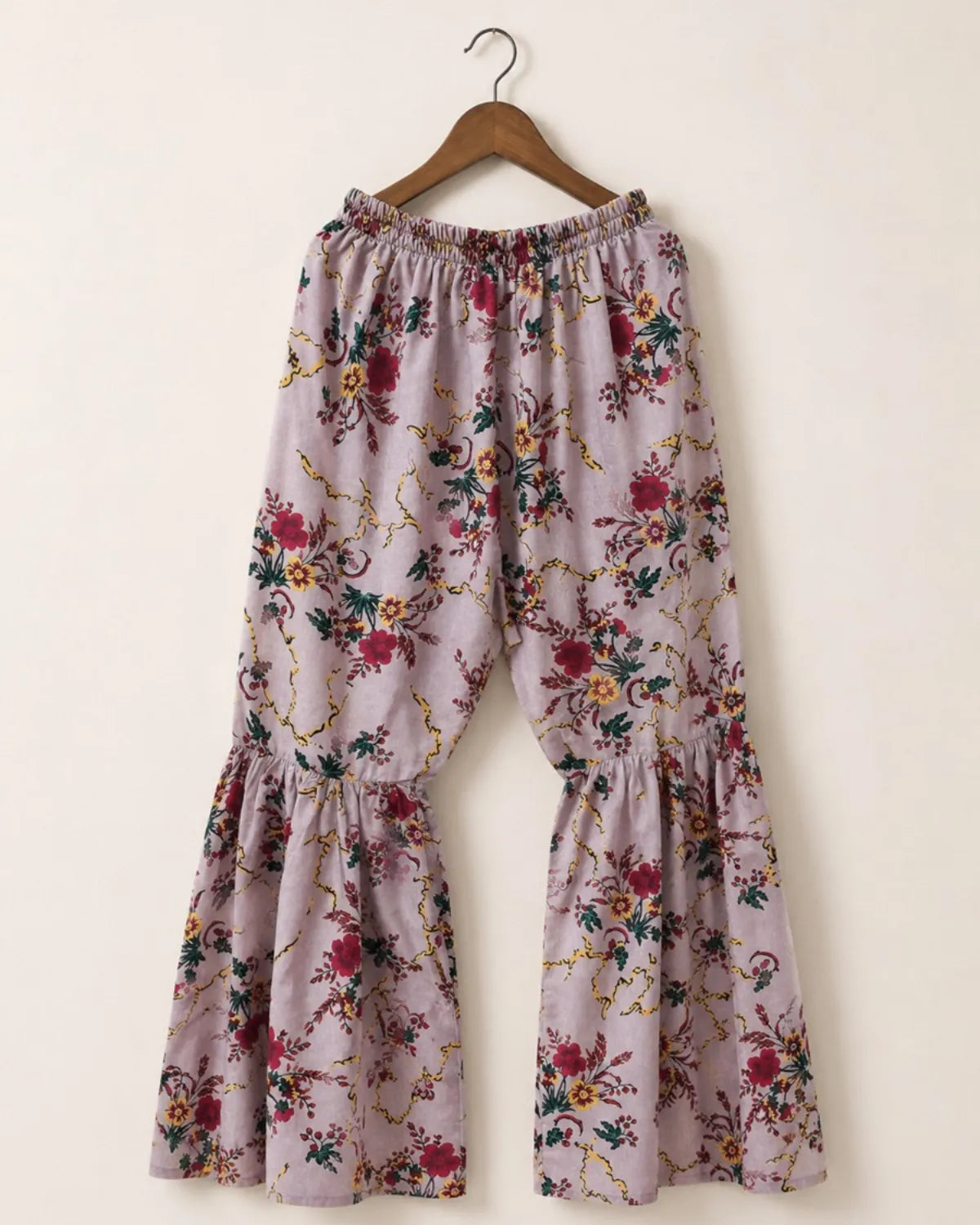 A pair of wide-leg, tiered pants with an elastic waistband, featuring a dusty lavender floral print with red, green, and gold details, hanging on a wooden hanger against a plain beige wall.