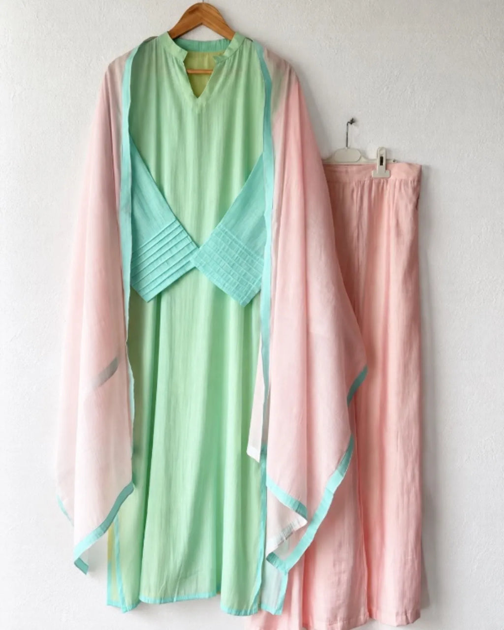 A light green and pink traditional-style dress set hanging on a wall, featuring a mint green tunic with a pleated wrap detail and a sheer pink outer layer, paired with matching pink pants.