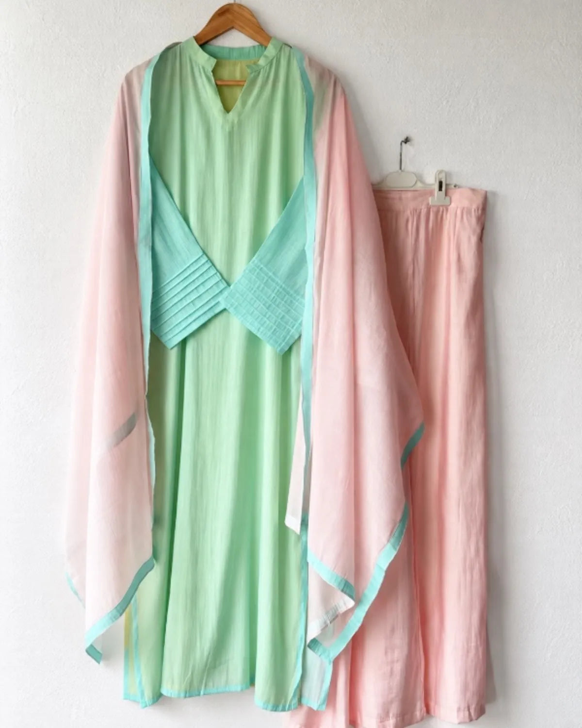 A light green and pink traditional-style dress set hanging on a wall, featuring a mint green tunic with a pleated wrap detail and a sheer pink outer layer, paired with matching pink pants.