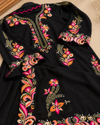 A black long-sleeve garment with vibrant floral and paisley embroidery in pink, orange, green, and gold, laid flat on a neutral beige surface.