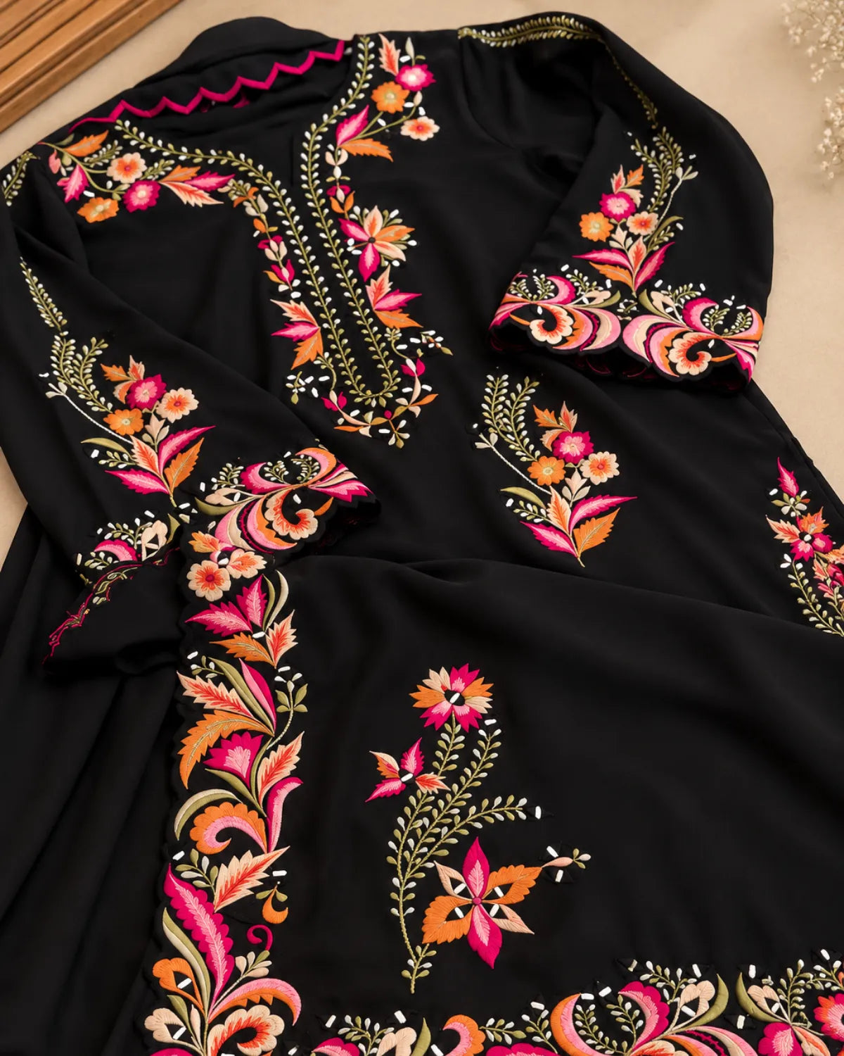 A black long-sleeve garment with vibrant floral and paisley embroidery in pink, orange, green, and gold, laid flat on a neutral beige surface.