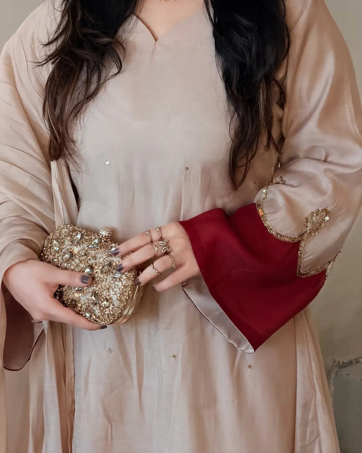 A close-up of a person wearing a beige traditional outfit with wide sleeves and a deep red contrast panel on the cuff, holding a small gold embellished clutch.