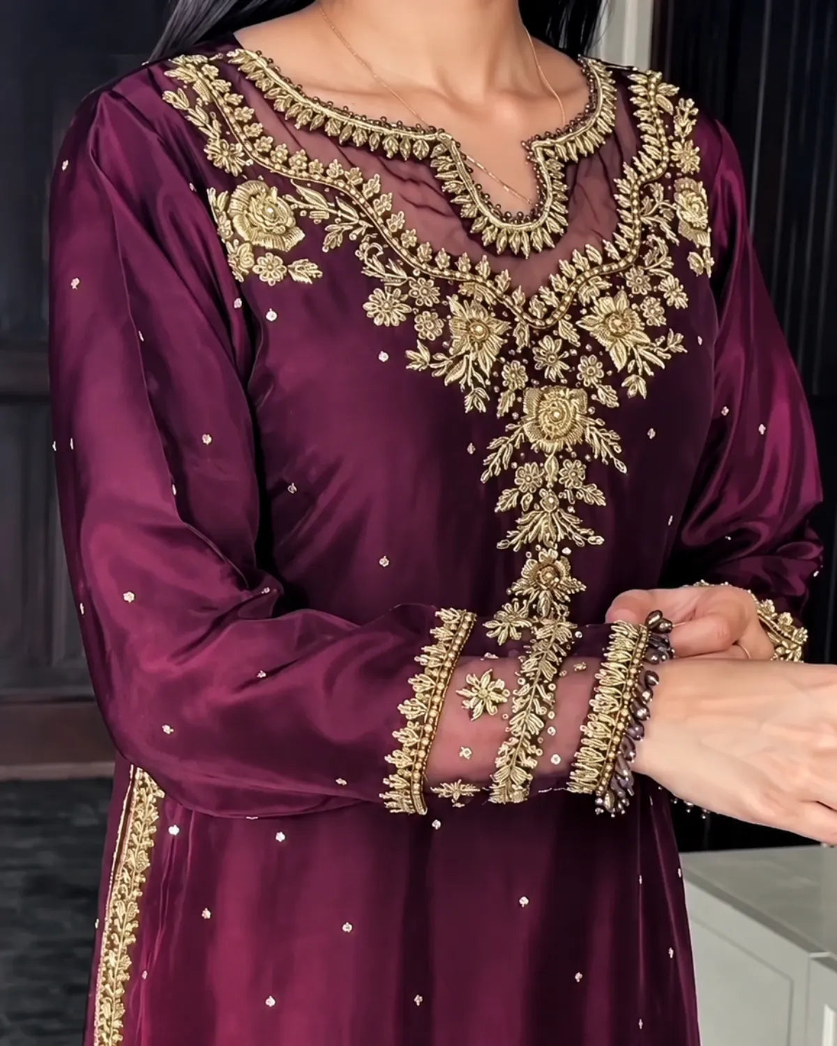 A close-up of a woman wearing a deep purple embroidered traditional outfit with ornate gold floral embroidery on the neckline, sleeves, and hem, along with sheer mesh panels.
