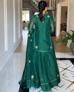 A woman viewed from behind wearing a traditional green anarkali-style dress with gold embroidery, standing in a modern hallway with white walls and tiled floors.