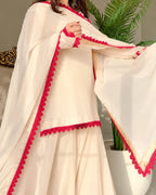 A cream-colored traditional outfit with bright red scalloped lace trim, modeled in an indoor setting with a potted plant in the background.
