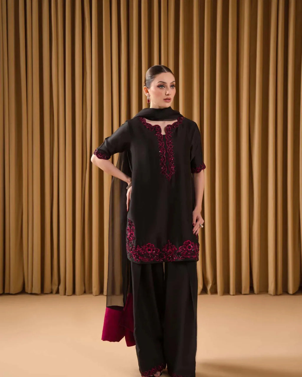 Woman wearing a black outfit with red embroidery against a beige curtain background