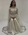 A woman seated in a studio wearing a formal white and silver embellished outfit with a sheer embroidered cape and wide flared pants.