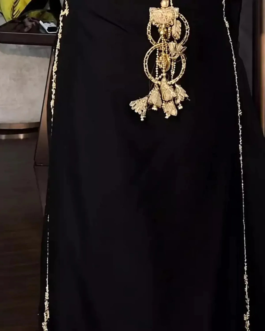 A close-up of a black garment with ornate gold embellishments, including a large dangling gold ornament at the center and thin gold chain-like details running down the sides.