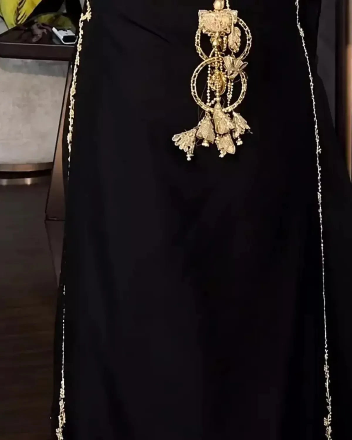 A close-up of a black garment with ornate gold embellishments, including a large dangling gold ornament at the center and thin gold chain-like details running down the sides.