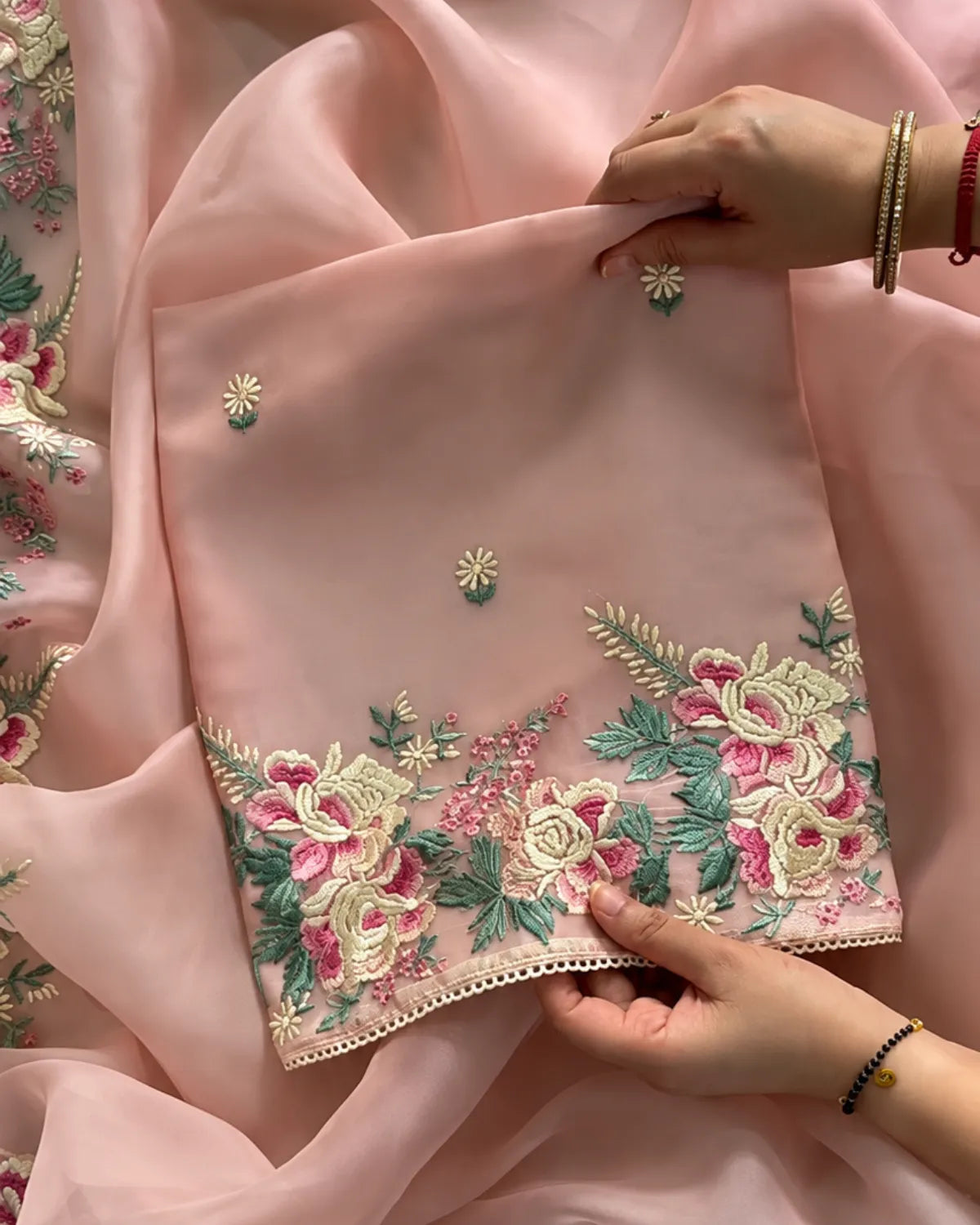 A close-up of a light pink embroidered fabric, likely a dupatta or scarf, held by hands to show its texture and detailed floral embroidery. The fabric features intricate embroidery with pink, green, and gold threads along a scalloped edge.