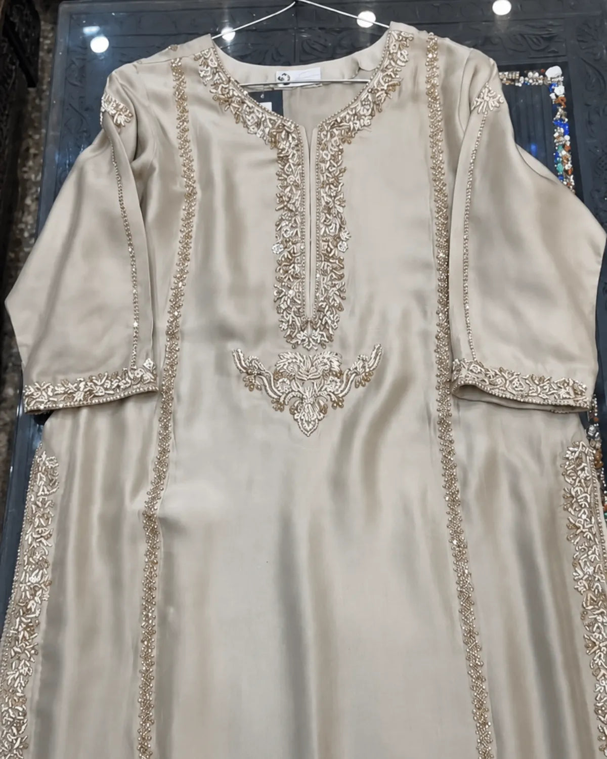 A beige satin kurta with intricate gold and silver embroidery, displayed on a hanger inside a glass display case.