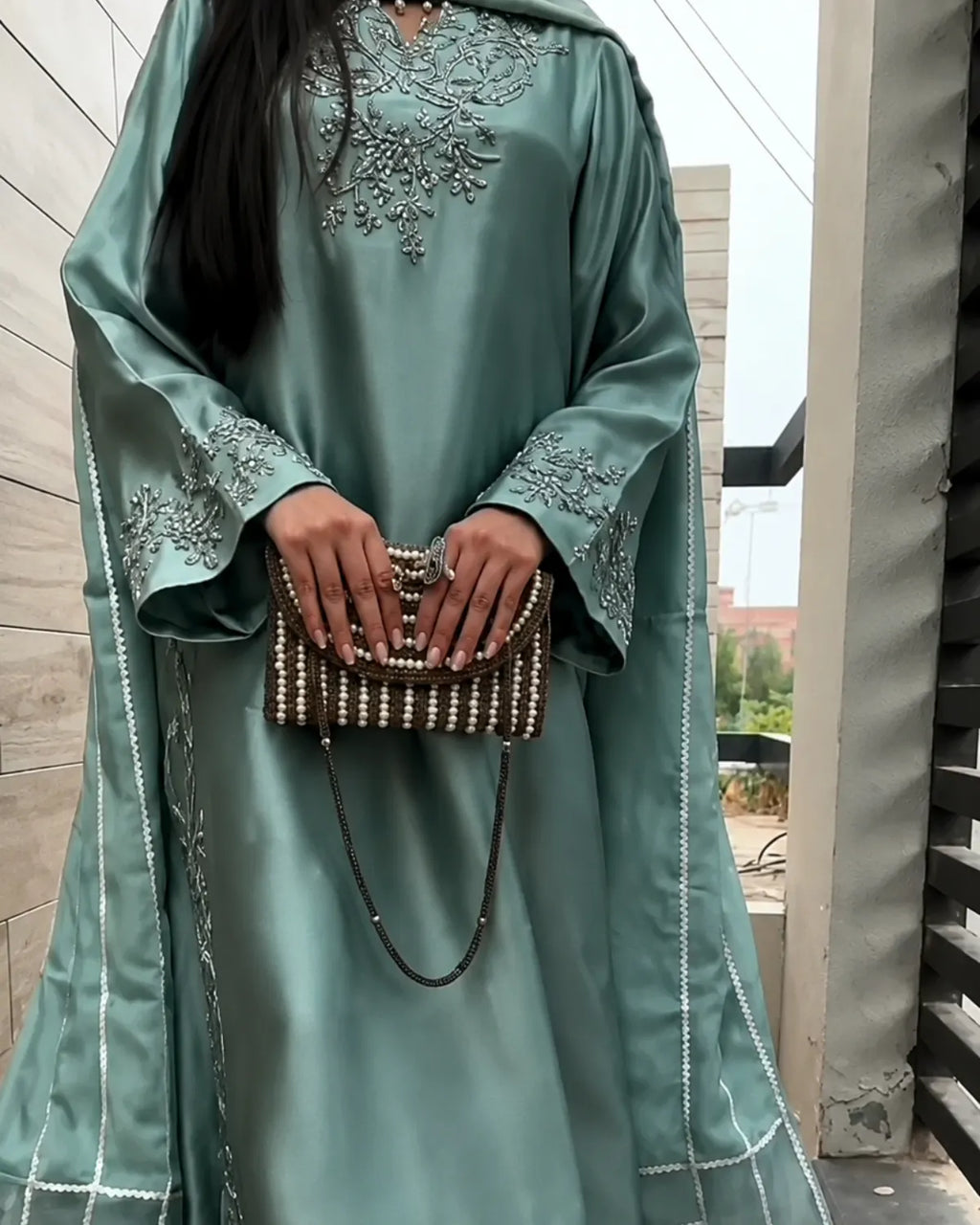 A woman wearing a flowing sage green abaya with silver embroidery on the chest and sleeves, holding a beaded brown and white clutch bag.