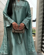 A woman wearing a flowing sage green abaya with silver embroidery on the chest and sleeves, holding a beaded brown and white clutch bag.