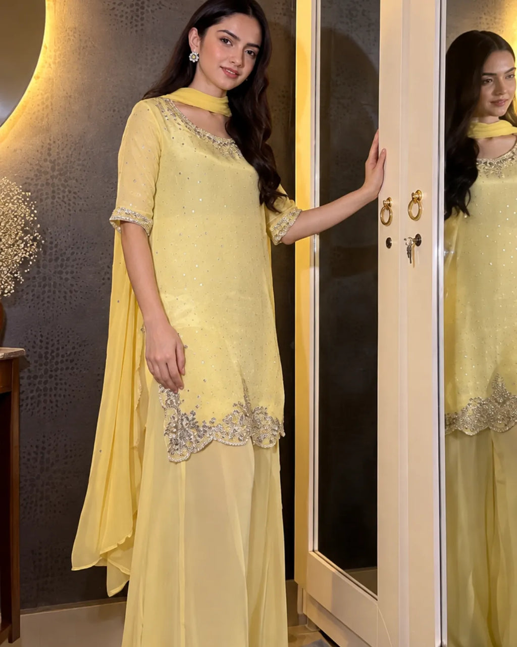 A woman models a light yellow ethnic kurta set with a matching dupatta and silver embroidered details, standing in front of a mirror in an indoor setting.