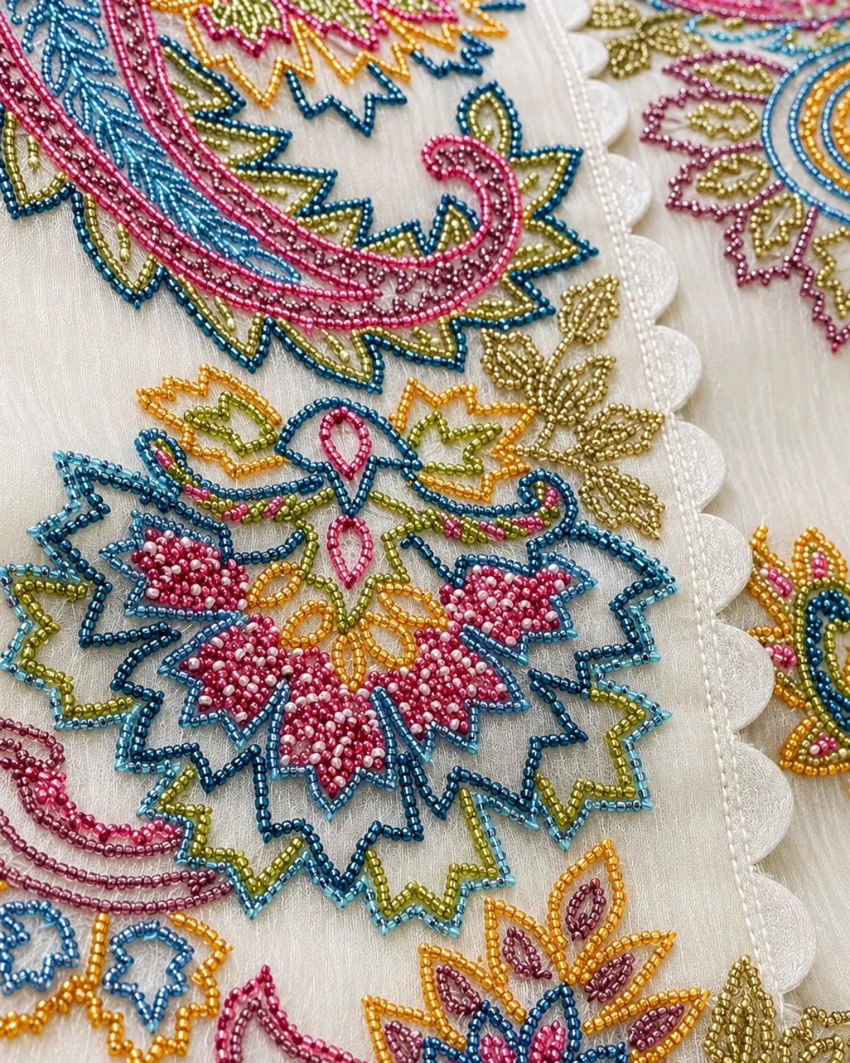 Close-up of an embroidered textile with a white scalloped lace trim and colorful beadwork in pink, blue, yellow, and green on a white base.