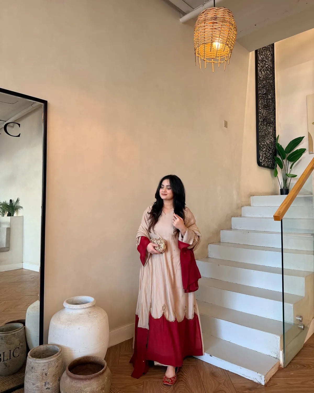 A woman wearing a beige and deep red tiered dress stands in a modern indoor setting with white stairs, a woven pendant light, and large ceramic pots.