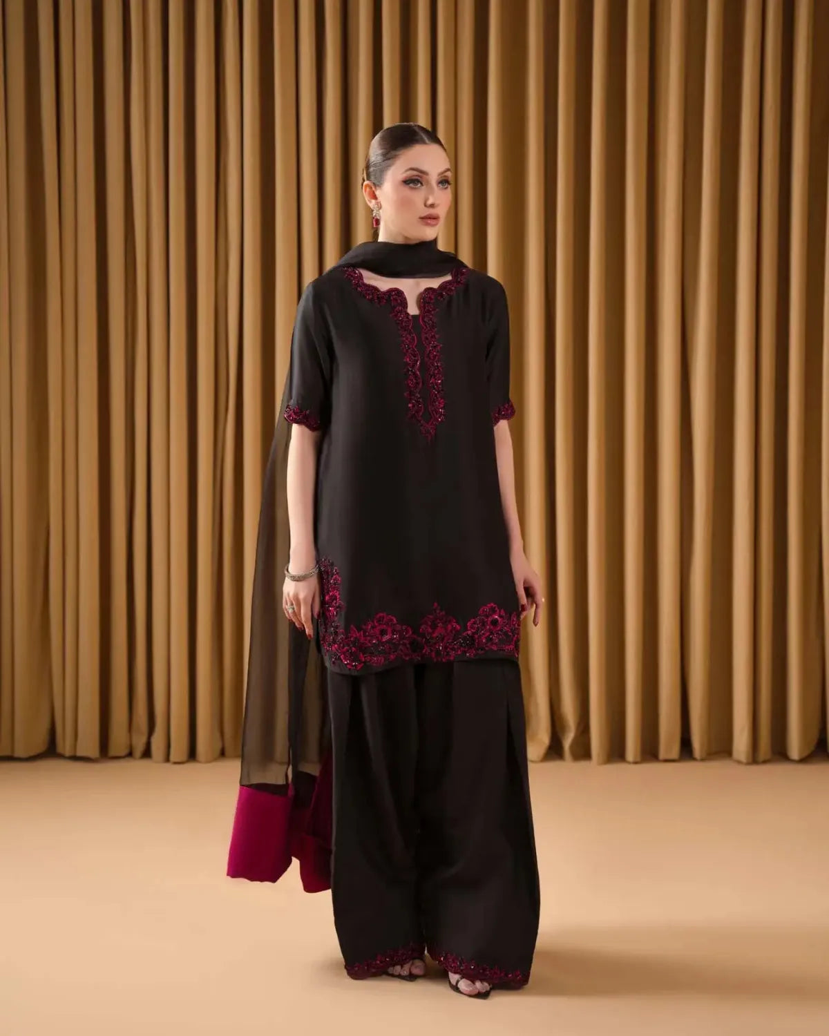 A woman modeling a black traditional outfit with deep red floral embroidery, consisting of a short-sleeved tunic, wide-leg pants, and a sheer black dupatta, set against a backdrop of gold curtains.