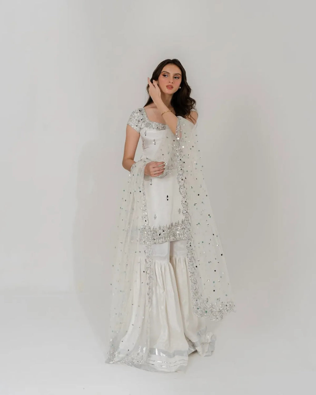 A woman modeling a white traditional outfit with a long sheer embellished dupatta, standing against a plain light gray studio background.