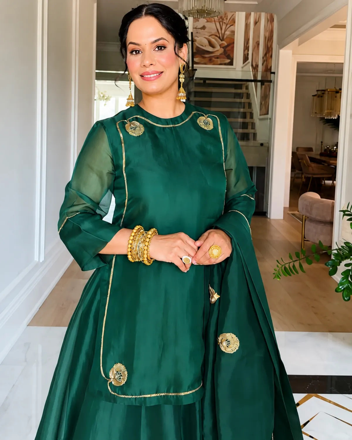 A woman wearing a dark green traditional outfit with gold embroidery and sheer sleeves, standing in a bright indoor hallway.
