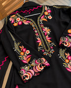 A black long-sleeve embroidered dress or tunic laid flat, featuring colorful floral and vine embroidery in pink, orange, yellow, and white around the neckline, cuffs, and hem.