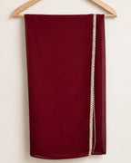 A deep burgundy red scarf draped over a wooden hanger against a plain off-white background, featuring a decorative cream-colored beaded border along one side.