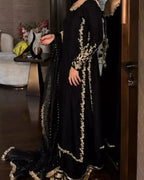 A woman wearing a black traditional outfit with ornate gold embroidery and a matching black dupatta, photographed indoors near a mirror and side table.