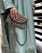 A close-up of a person wearing a light sage green embroidered traditional outfit and holding a brown and cream beaded clutch bag with a braided strap.