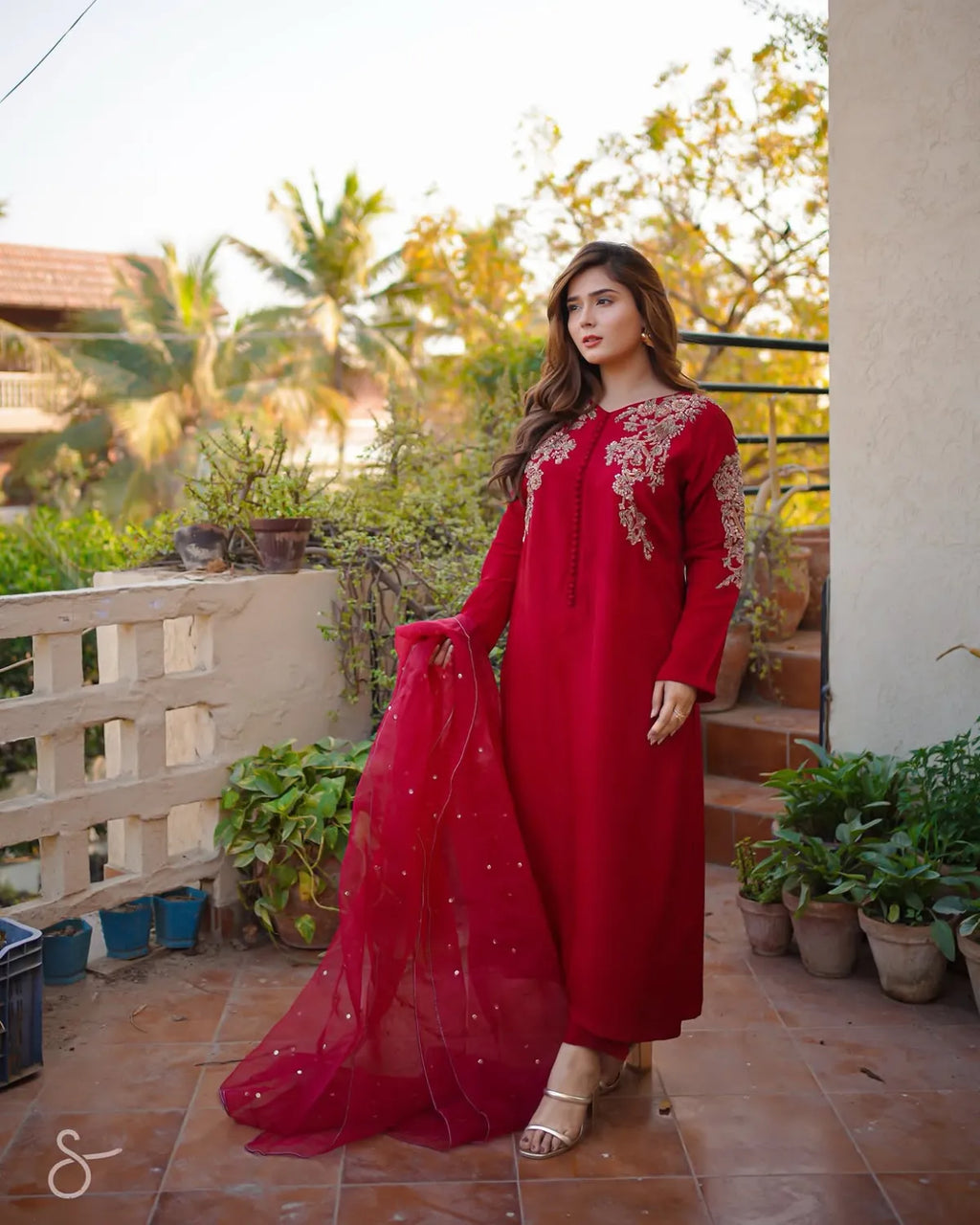A woman wearing a long-sleeved red embroidered kurta with a matching sheer dupatta, standing on an outdoor balcony with plants and a tiled floor.