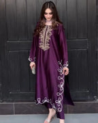 A woman wearing a long, flowing purple kaftan dress with white and gold embroidery, standing in front of a dark paneled door.