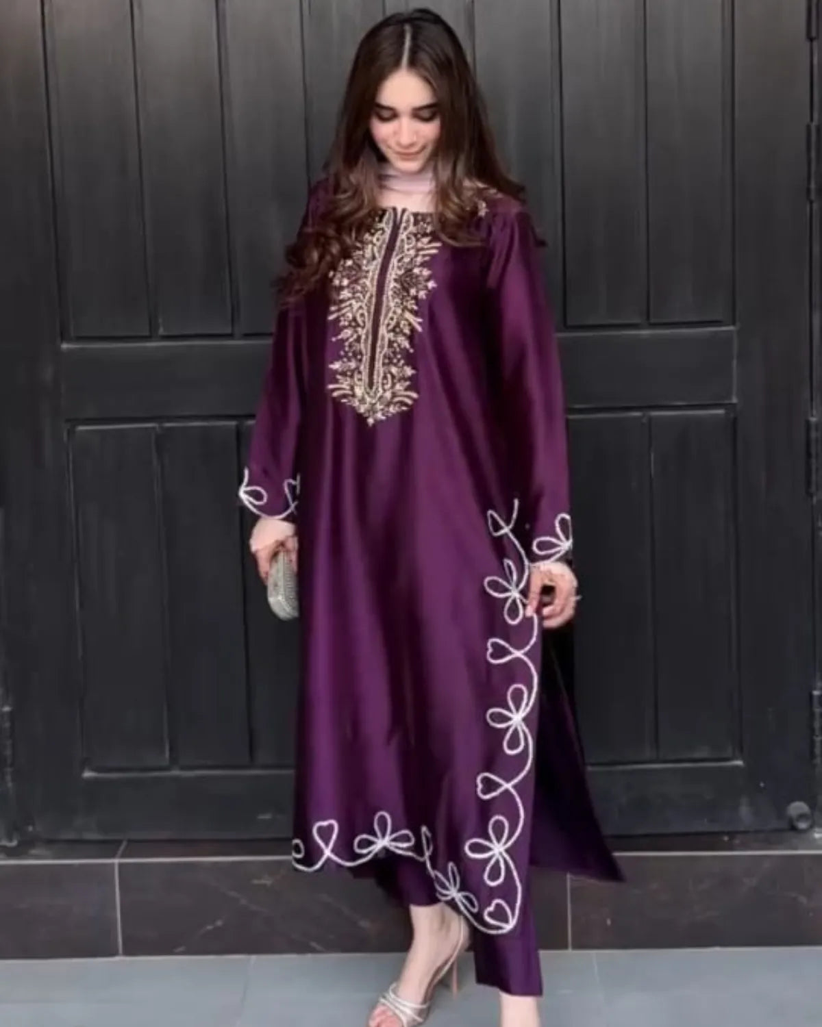 A woman wearing a long, flowing purple kaftan dress with white and gold embroidery, standing in front of a dark paneled door.