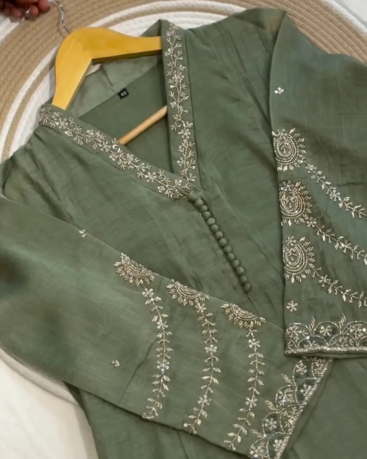 An olive green traditional tunic with intricate silver and white floral embroidery, displayed on a wooden hanger over a woven mat background.
