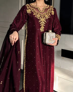 A woman wearing a deep maroon traditional outfit with ornate gold embroidery around the neckline and along the border, holding a silver embellished clutch. The background shows a modern interior with white walls and dark panels.