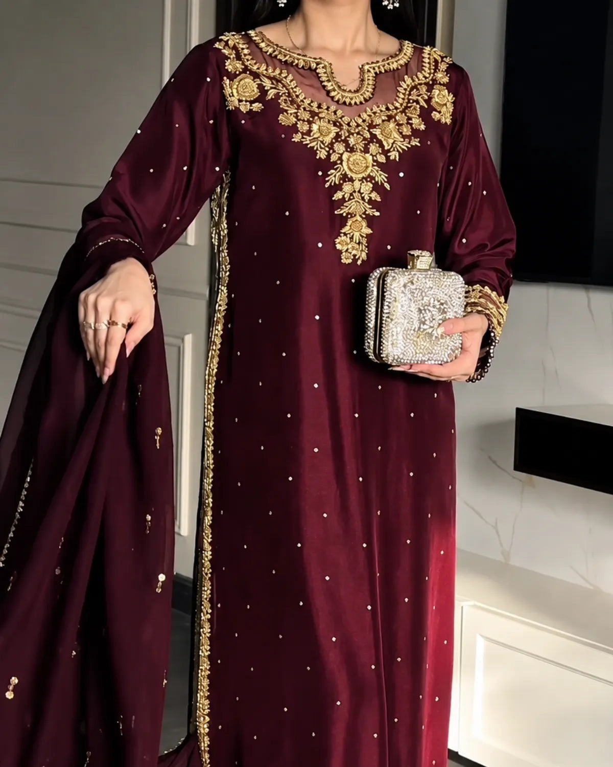 A woman wearing a deep maroon traditional outfit with ornate gold embroidery around the neckline and along the border, holding a silver embellished clutch. The background shows a modern interior with white walls and dark panels.