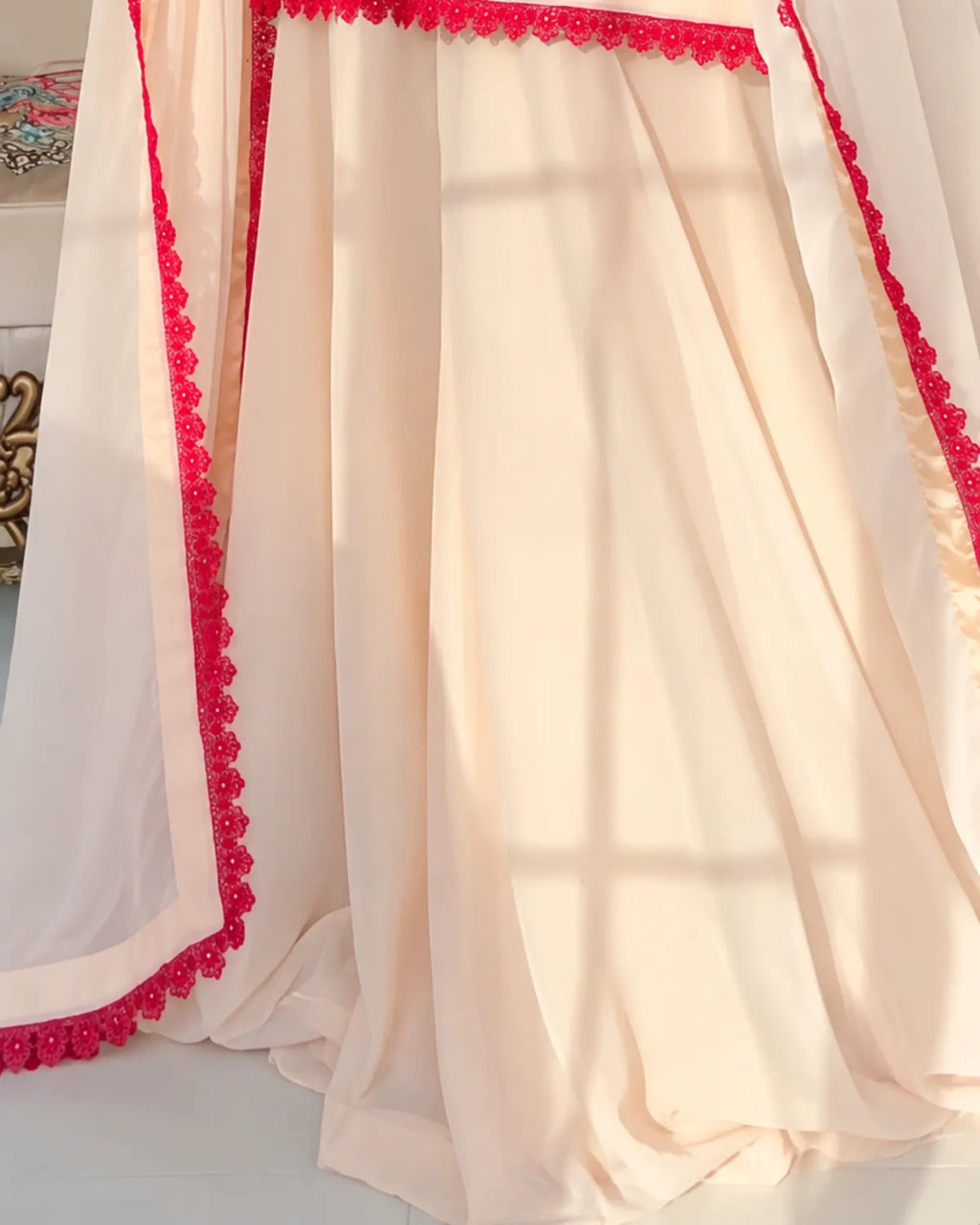 A draped cream-colored sheer fabric with bright pink floral lace trim along the edges, shown in soft folds with warm sunlight filtering through.