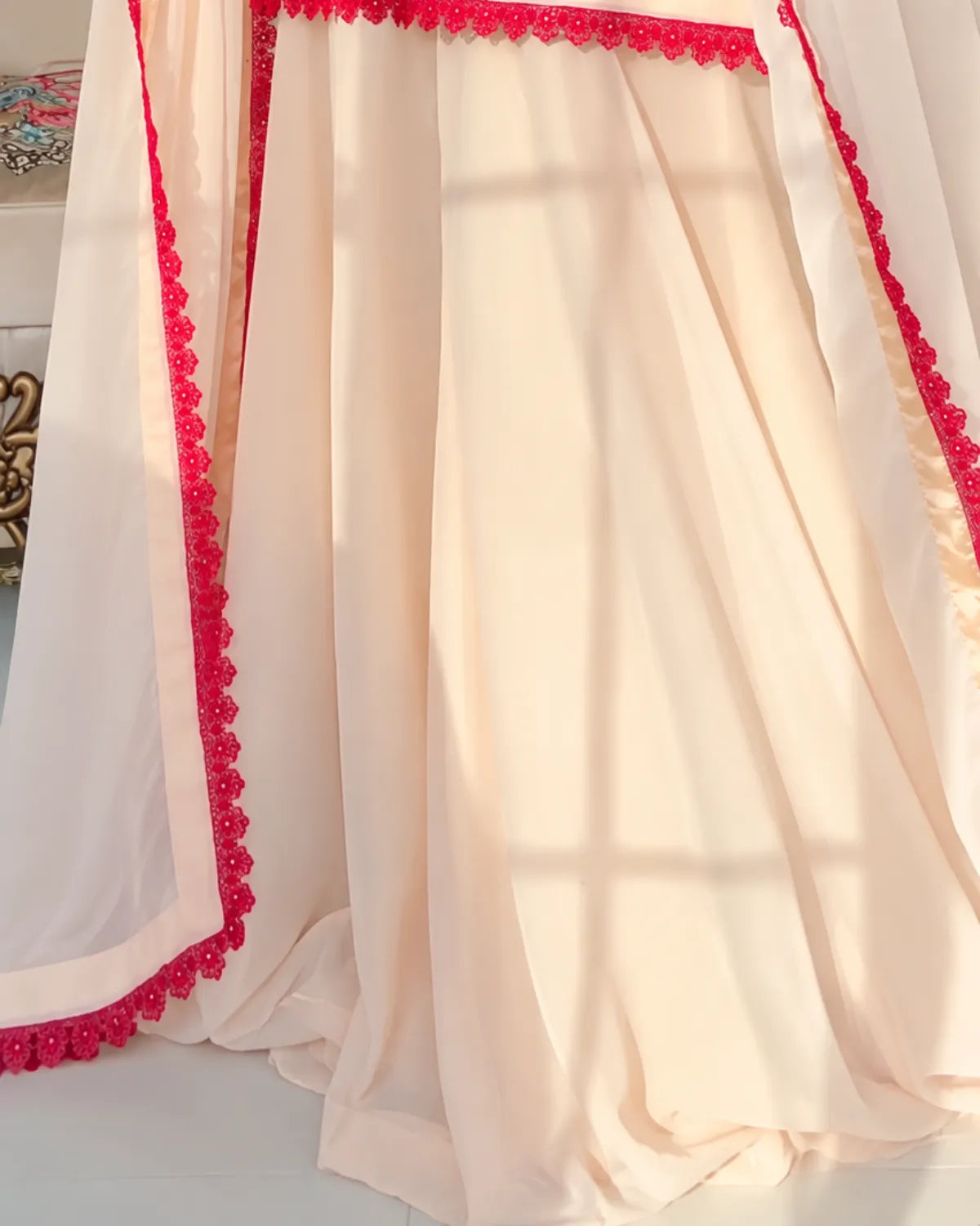 A draped cream-colored sheer fabric with bright pink floral lace trim along the edges, shown in soft folds with warm sunlight filtering through.